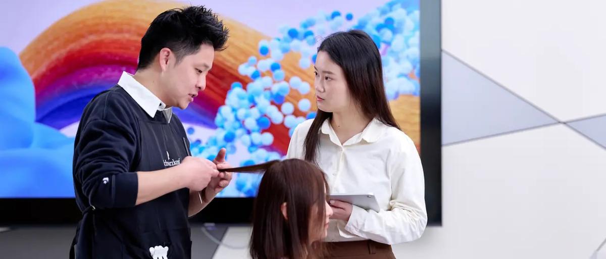 Two people talking as one looks at a seated persons hair.