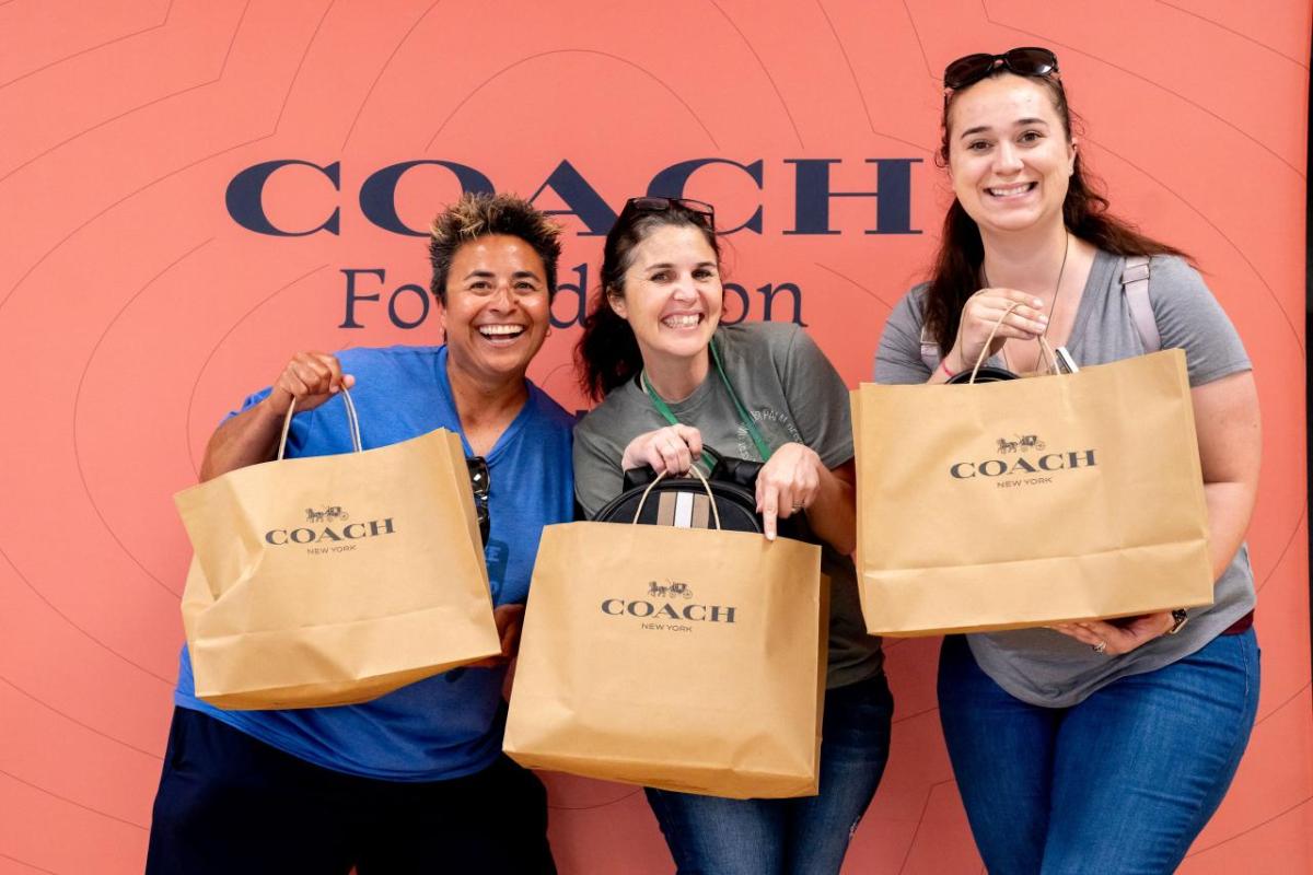 Three woman stand together holding Coach shopping bags with in front of a salmon pink colored backdrop with the Coach logo