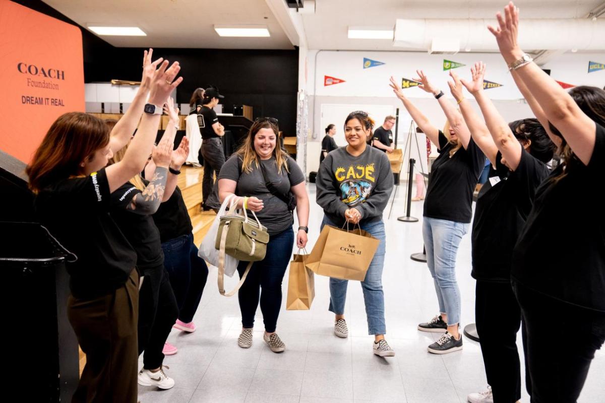 People standing in two lines with their arms raised while two women holding Coach shopping bags walk down the middle