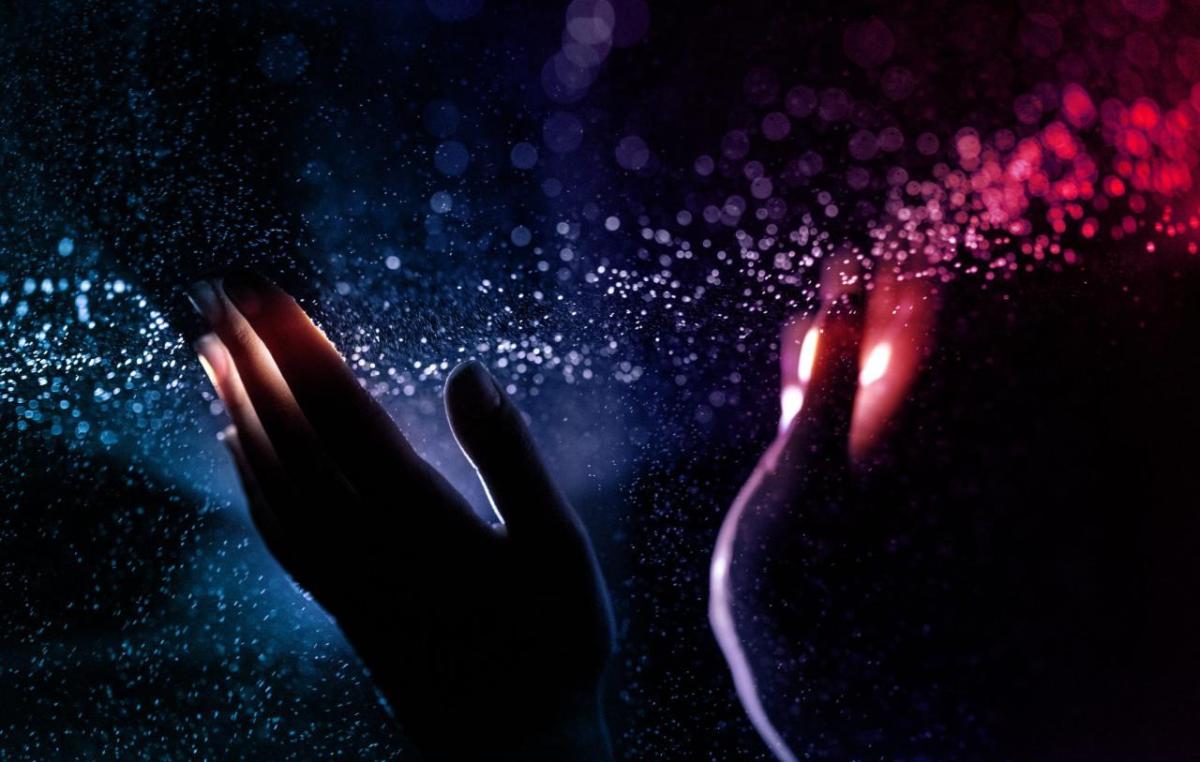 Two hands upheld in a dark space. A narrow band of colorful light and particles streaming across the finger tips.