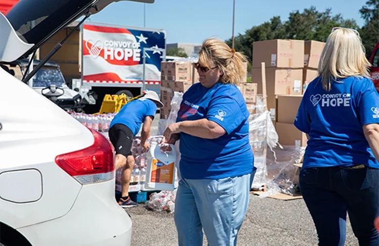 Volunteers loading the trunk of a vehicle. "Convoy of Hope" sign behind them and stacks of boxes.