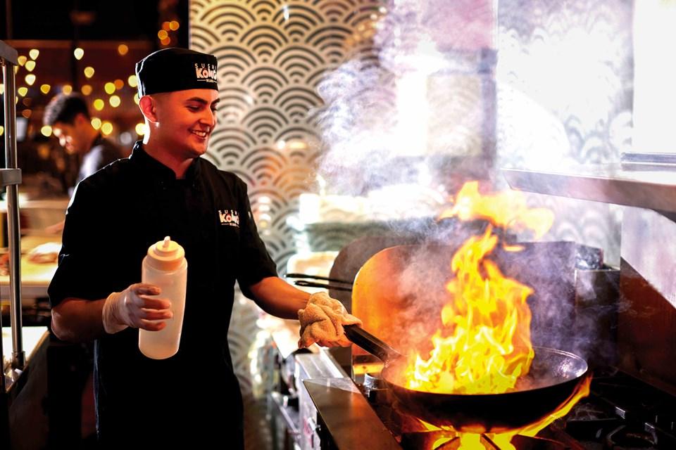 A chef smiling looking at a flaming wok on a stove.