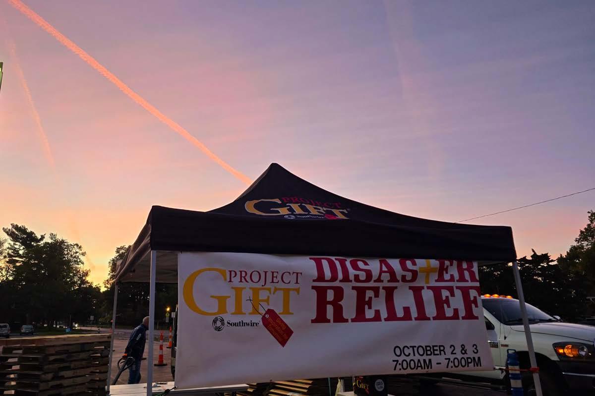 A pop-up canopy with Project Gift banner hanging on the front, Outside in a parking lot. A setting sun in the background.