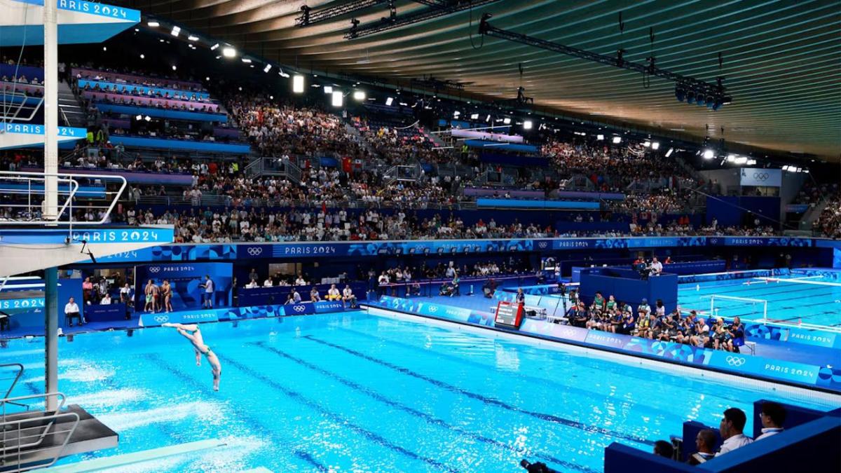 Two people diving into a large pool. Spectators in the stands.