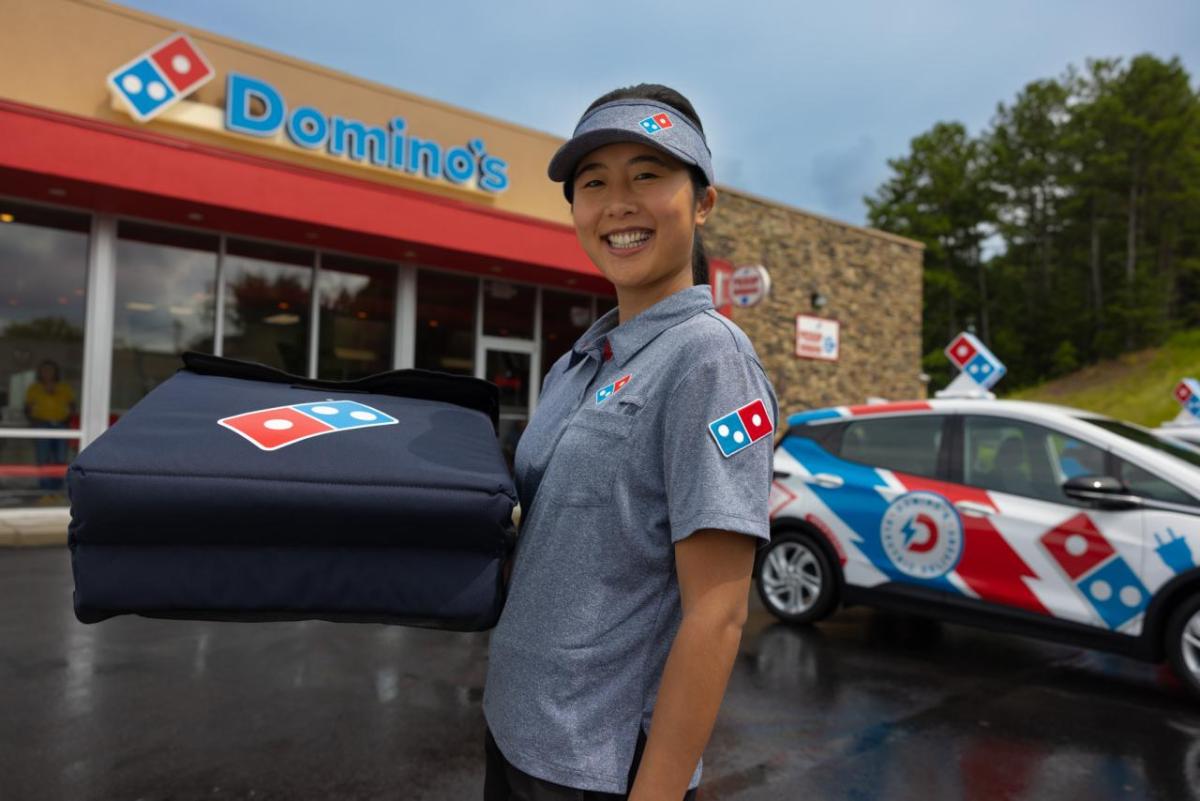 A smiling person in Domino's uniform, holding a pizza delivery box. A delivery car and Domino's store behind them.