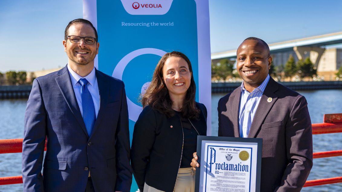 Three people standing next to a waterway, one holding a proclamation. 