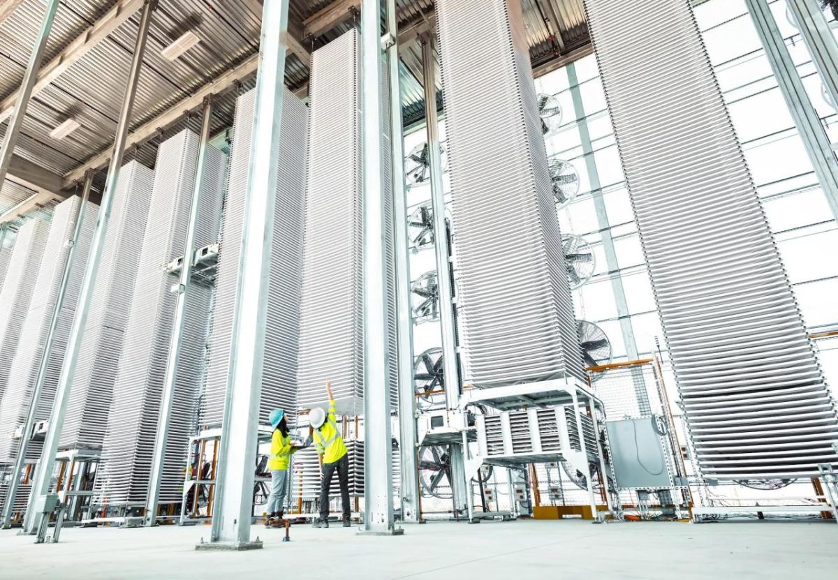 Two people in high-vis vests looking at a tall stack of metal sheets in a row of other stacks.