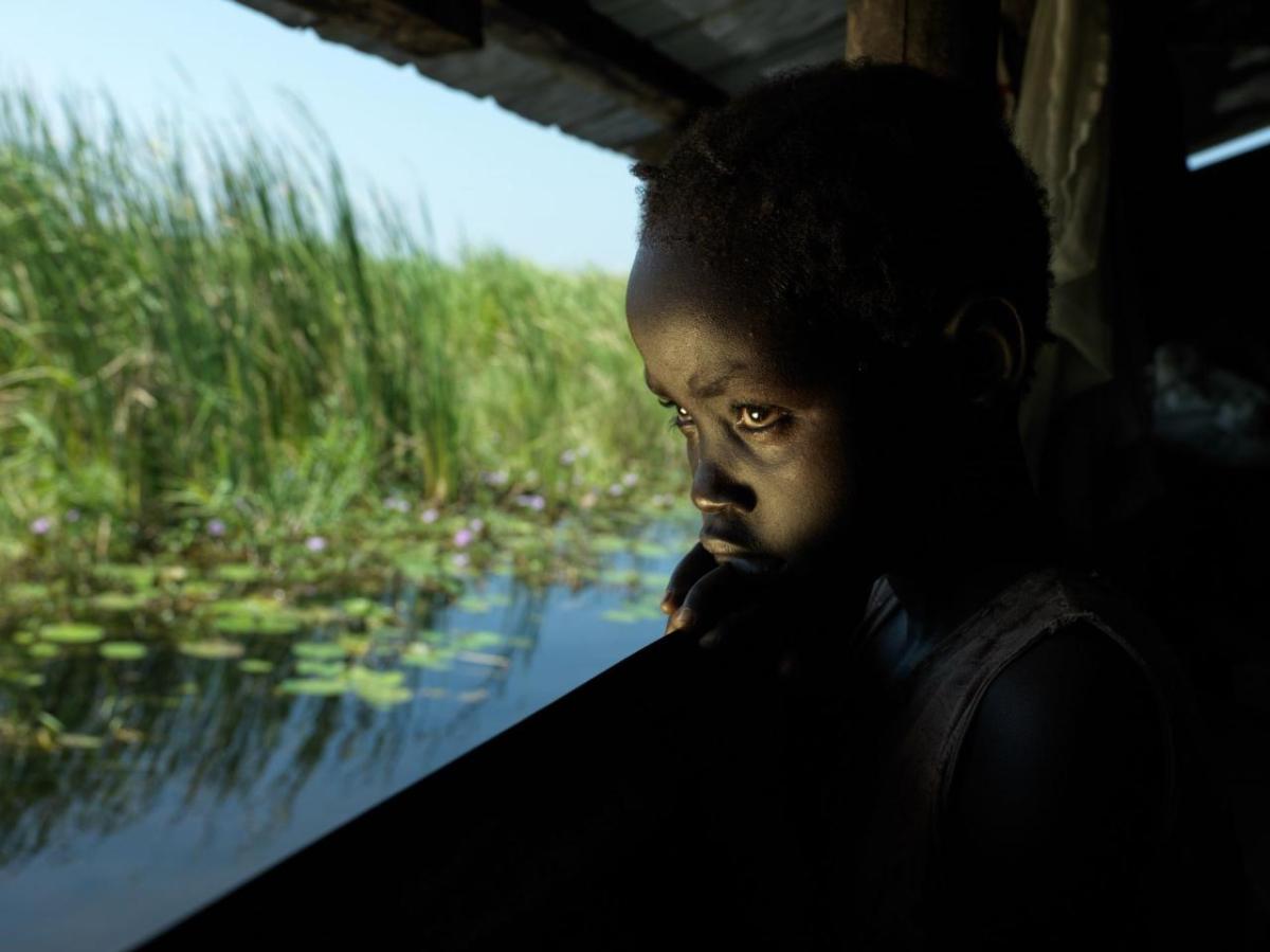 Ten-year-old, Nyma waits for her mother to return home from serving tea to the local authority. They both live in an abandoned classroom in the school she used to attend before it closed due to the floods. Using locally sourced grass, they built a floating pontoon on top of the concrete classroom floor to mitigate the rising flood water. Nyma gets anxious when her mother is away and misses her old home.