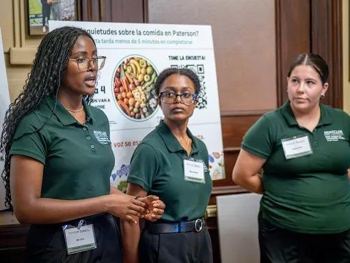 Three people in green shirts presenting. A display board behind them.