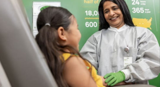 A smiling adult in protective gear looking at a child, seated.