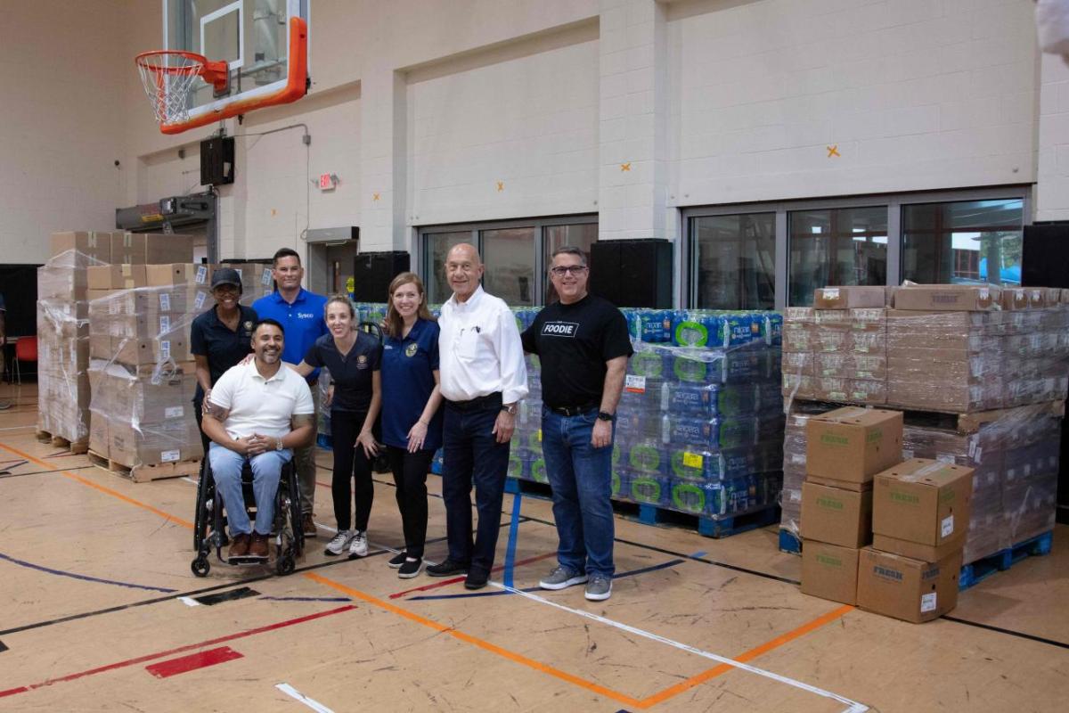 People posed in front of pallets of boxes of food, water and supplies in a gymnasium.