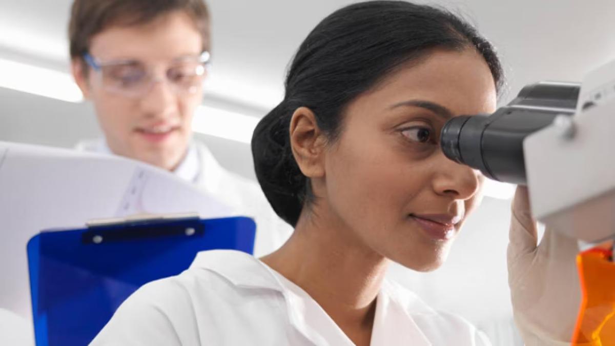A person looking into a microscope, another looking on with a clipboard.