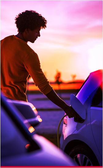 A person holding a plug to an electric vehicle port. A setting sun in the distance.