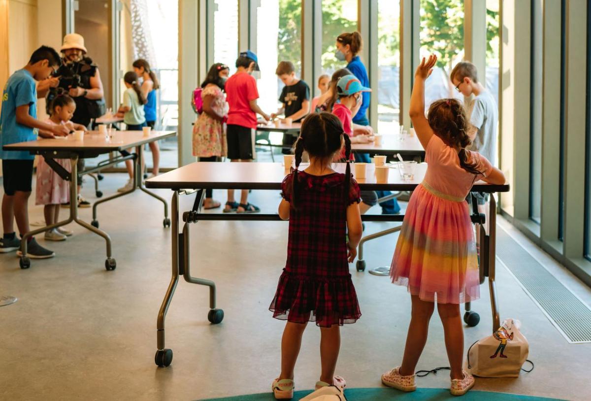 Kids standing at tables, one with hand raised.