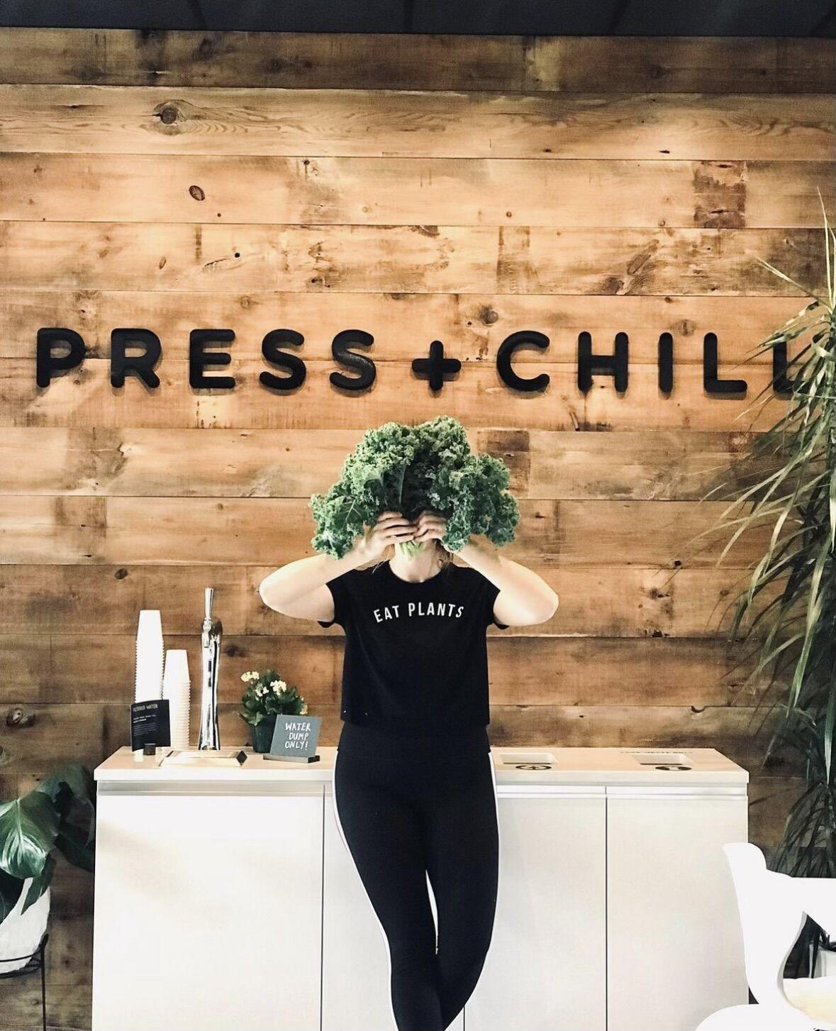 Kimberly Hoffmann holding a plant in front of her face. A sign on the wall behind them "Press + Chill".