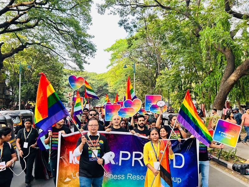 People marching in a parade waving rainbow flags and "Kohler Proud" banner.