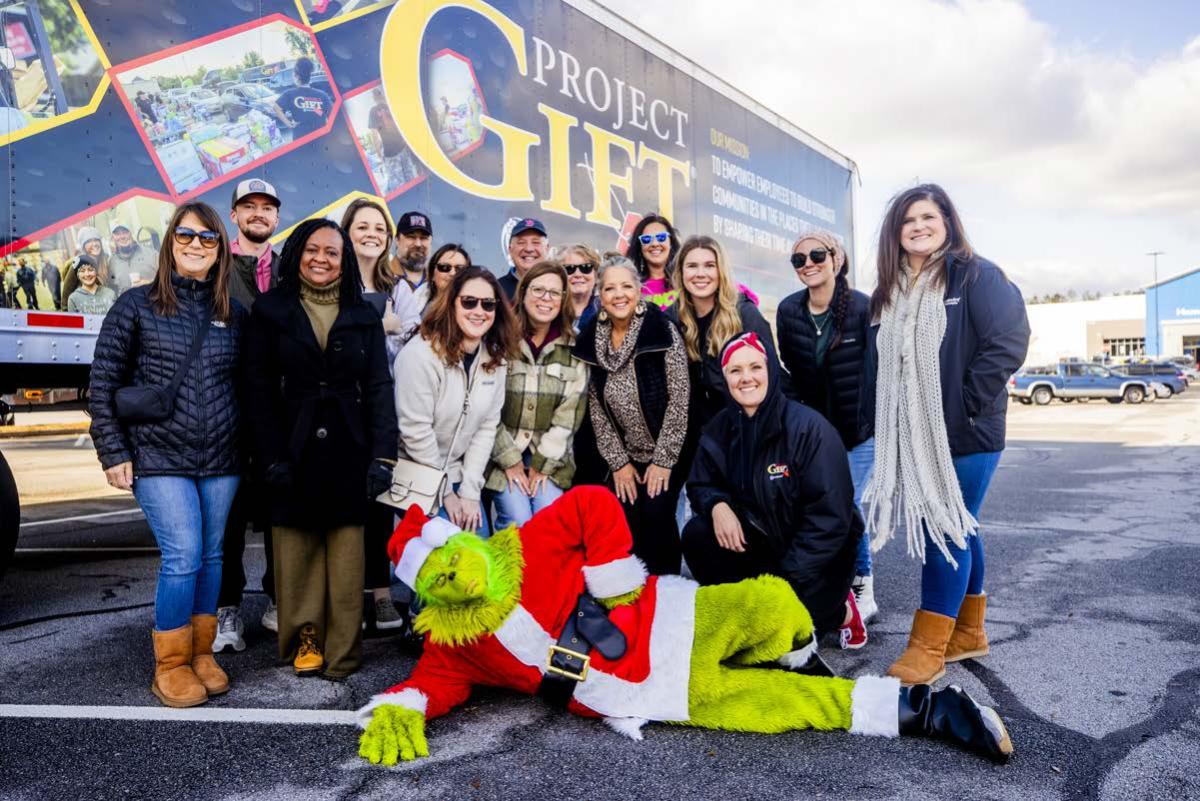 A group posed in front of a semi truck. A person dressed as the Grinch laid out in front of them.