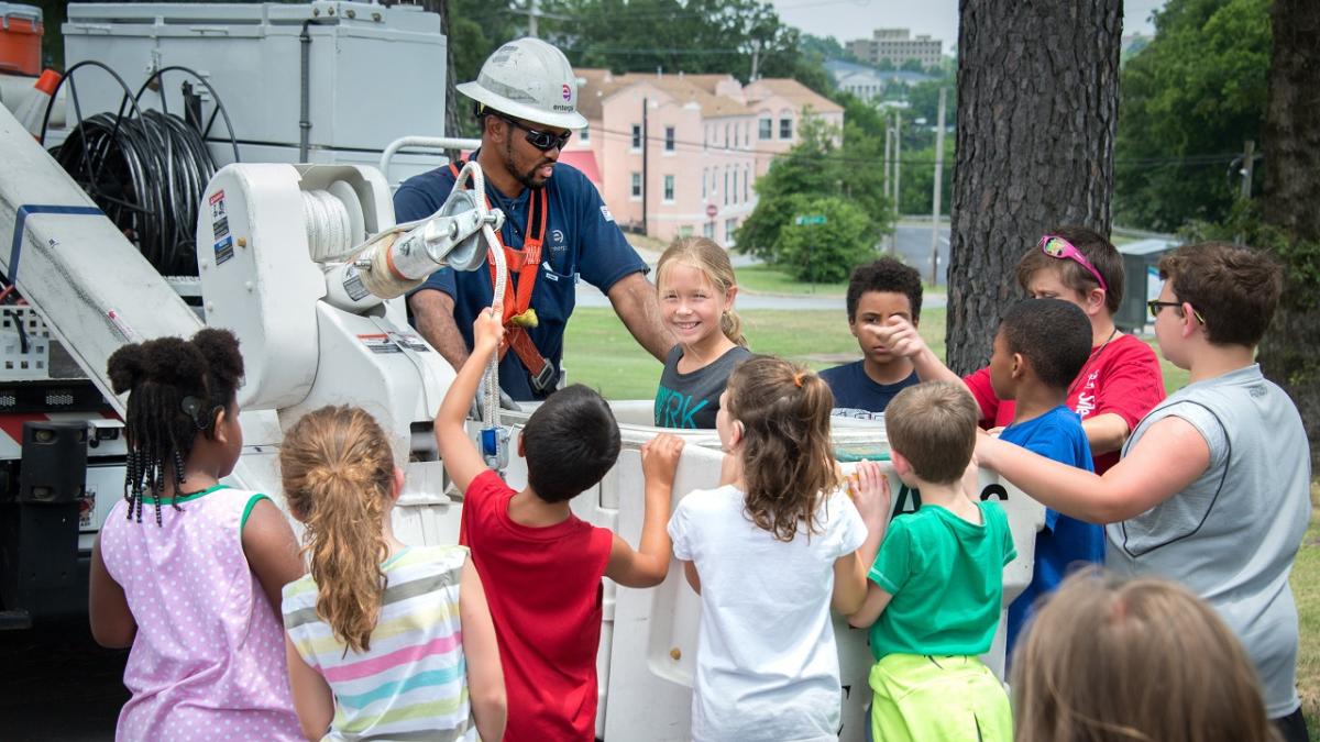 A person in safety gear helping a group of kids surrounding a bucket truck.