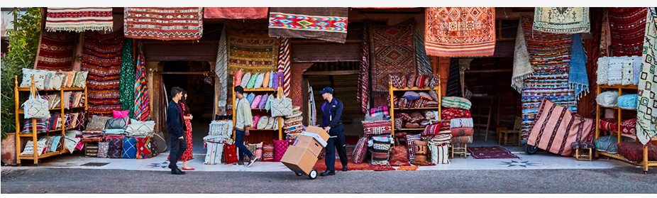 A person delivering boxes at a shop with colorful textiles hanging.