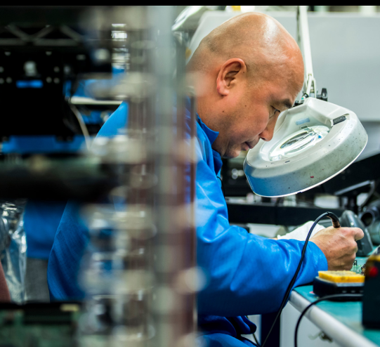 A person seated using a magnifying glass and soldering iron.