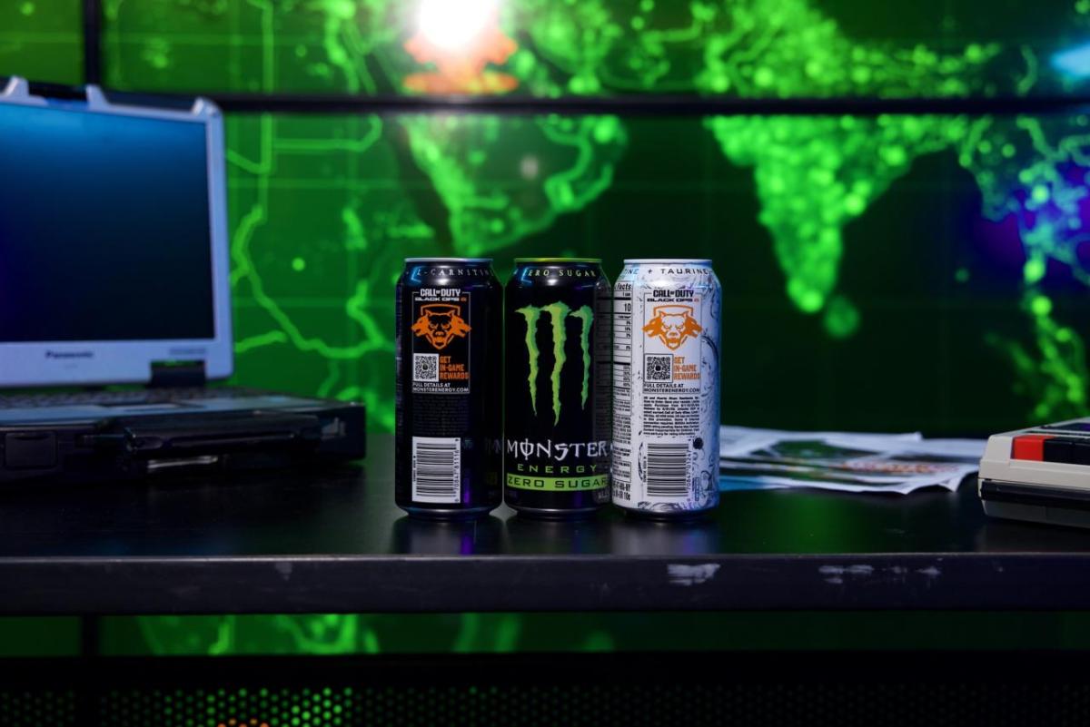 Three cans of Monster beverages on a black desktop in a dark room with a green-lit map on the wall behind them.