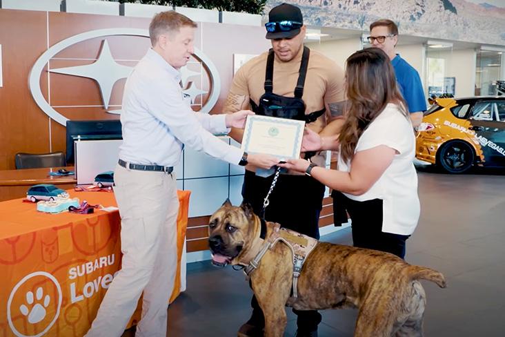 A person handing over a certificate to a couple who are stood with a service dog 