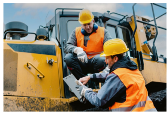 Two people talking, one crouching on a large construction vehicle, the other pointing to something they are holding.