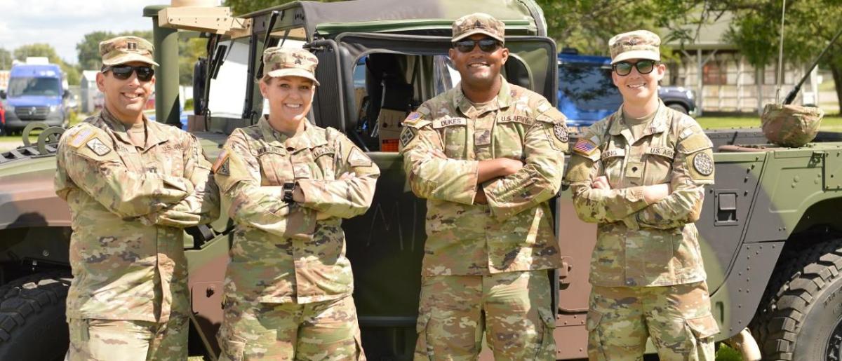 Four people posed with arms crossed in military uniforms. A military vehicle behind them.