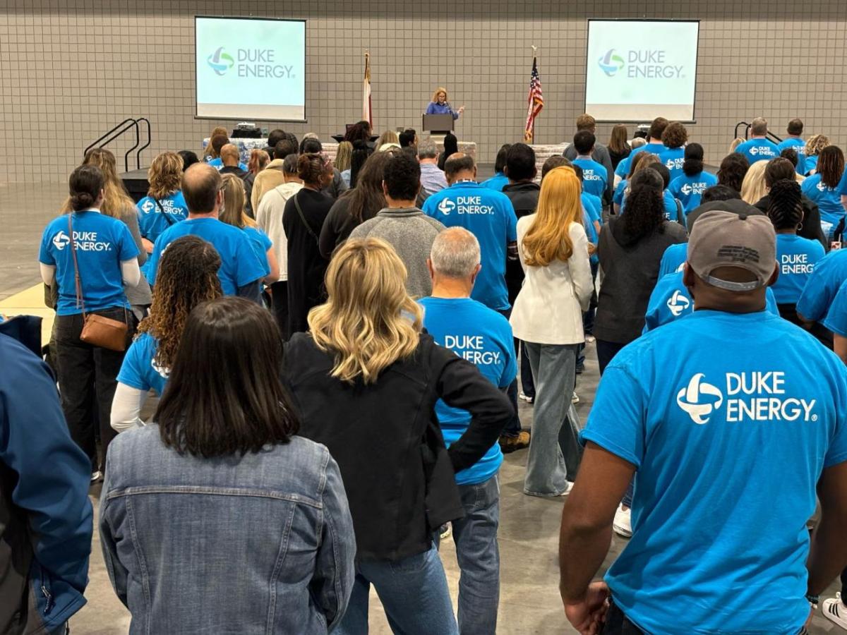 A person speaking on a stage to a group of volunteers and others standing in an open area.