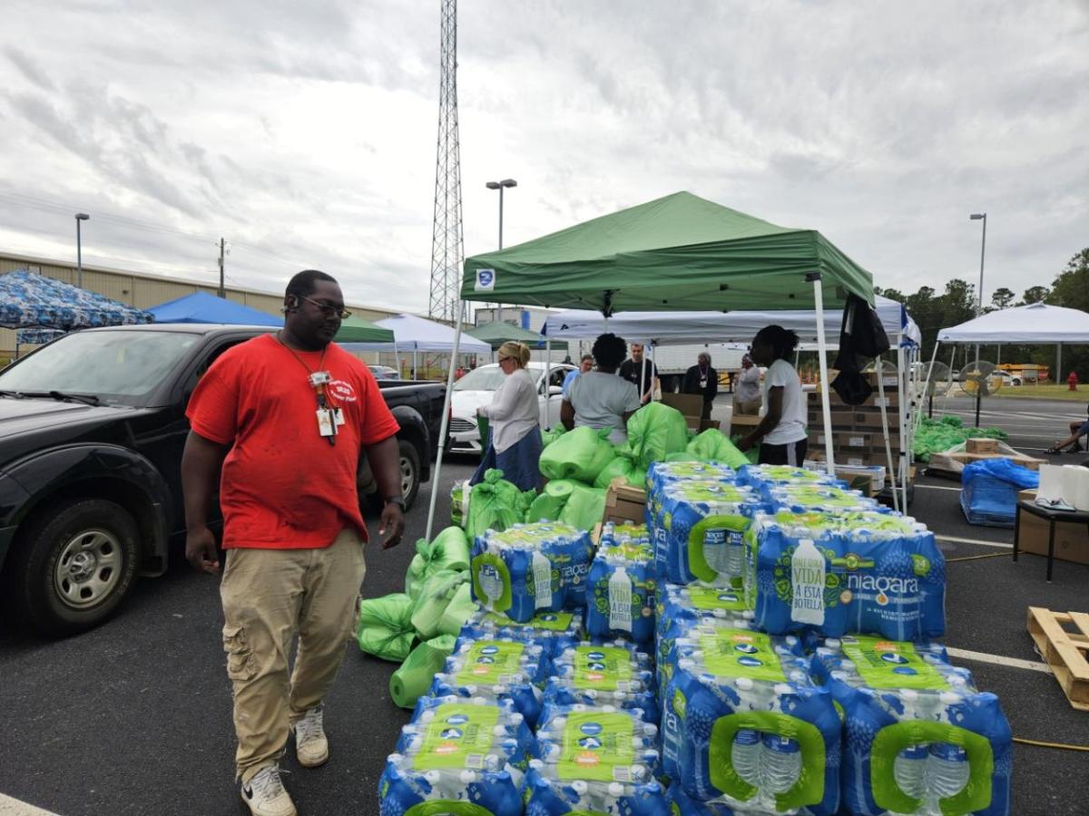 Stacks of wrapped water bottles outside, vehicles parked nearby