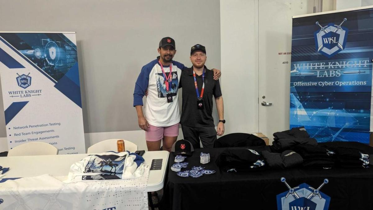 Two people posed behind tables, signs to the sides "White Knight Labs"