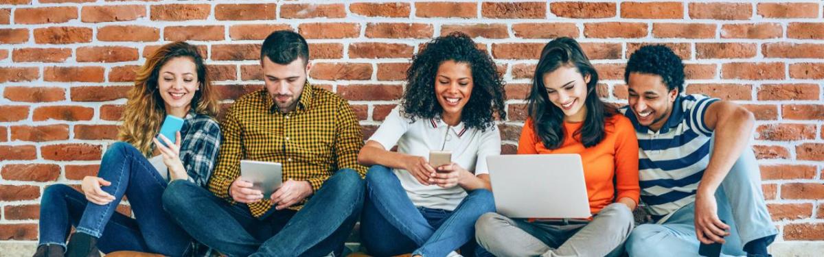 A group seated against a brick wall. Some smiling and looking at an open laptop or other devices.