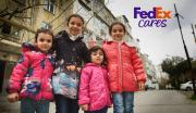 Four children posed close together, smiling outside residential buildings. "FedEx Cares" in the corner.